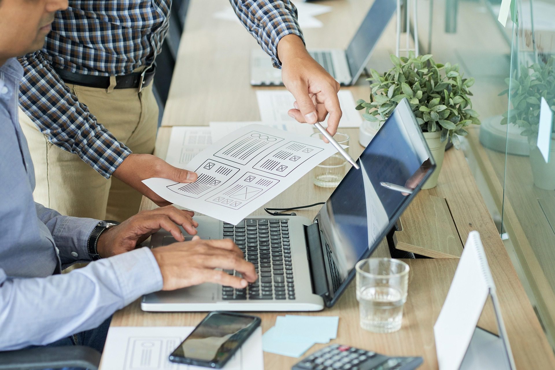 Two coworkers collaborating at a desk, one typing on a laptop while the other points at the screen and holds a paper with interface wireframe sketches.
